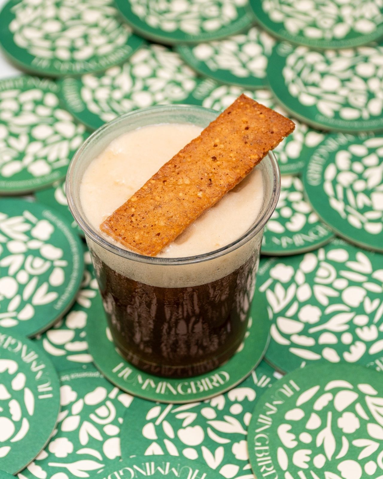 An iced pour with a cracker garnish, on a green patterned coaster