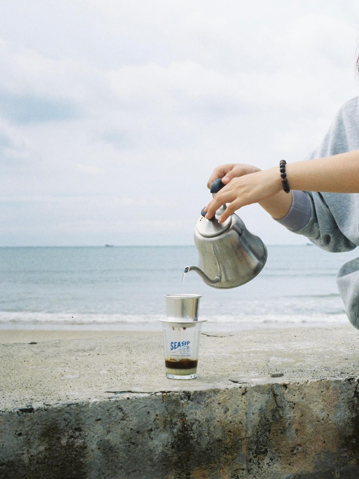 A branded Sea Sip iced coffee cup on the beach, next to a pour-over in a glass server