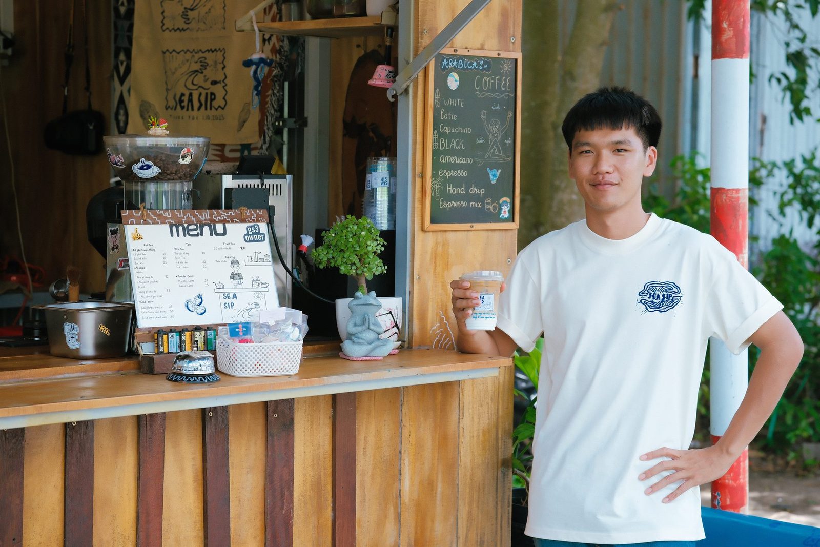 Quang standing by the counter, drink in hand, the menu board behind him