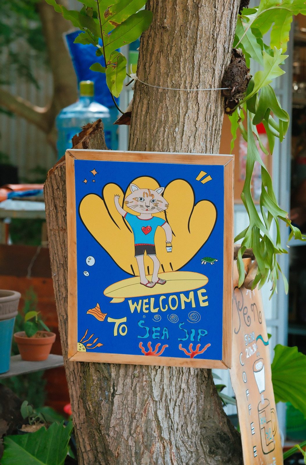 A hand-painted wooden sign of a surfing cat reading 'Welcome to Sea Sip', mounted on a tree trunk